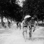 Two cyclists riding on the dusty road at the Tour de France in 1923, one of them is holding an umbrella