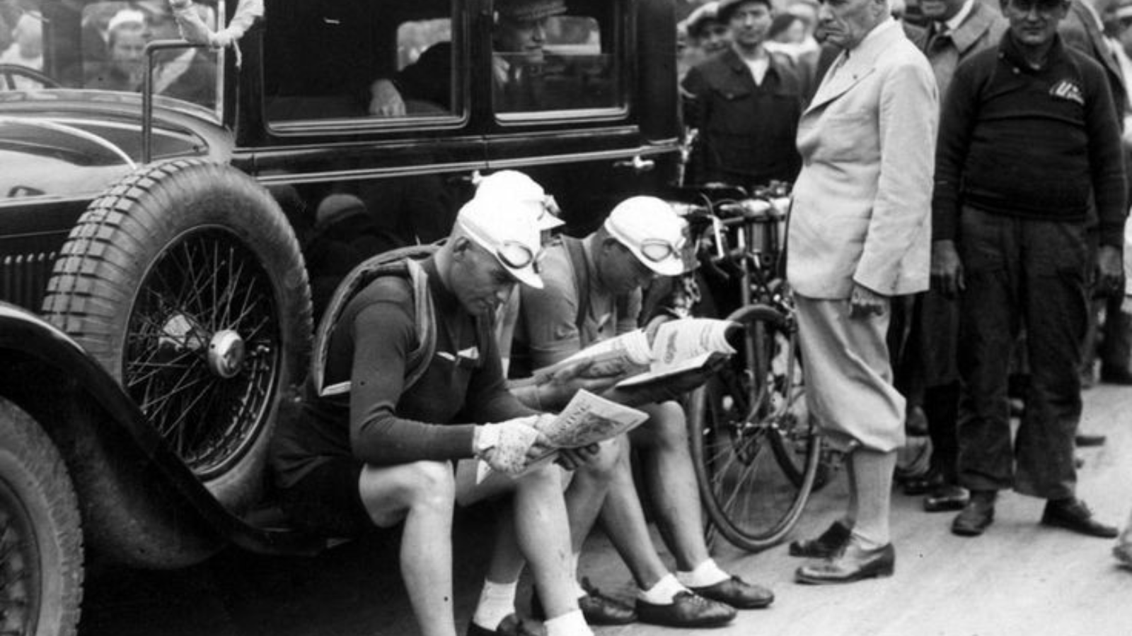 Black and white vintage picture about cyclists sitting at the side of a car and reading newspaper at the Tour de France 1931