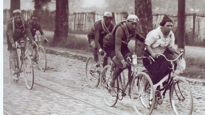 A woman, possibly accidentally riding her bicycle surrounded by some professional riders at the Tour de France in the 1920s