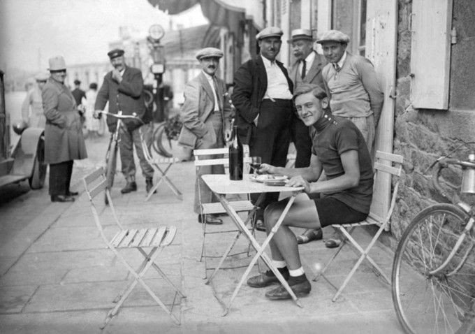 Austrian cyclist Max Bulla posing for the camera at the Tour de France in 1930. He is sitting at a small desk. Curious people are surrounding him, partially also looking into the camera. 